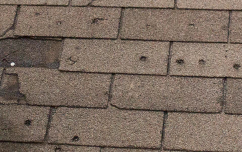 A close-up view of a roof with asphalt shingles in Madison, WI showing significant wear and damage. Some shingles are cracked and have dark patches, along with visible nail holes and spots where shingles are missing. This roof is a prime candidate for exterior renovations or complete replacement.