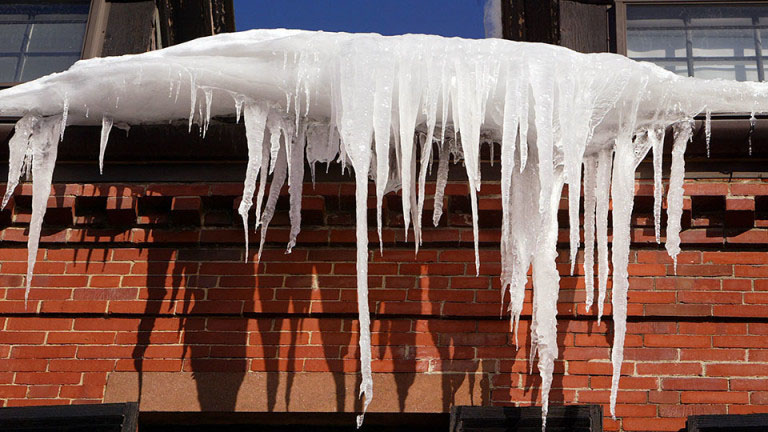 Large icicles hang from a snow-covered roof in Madison, WI, casting distinct shadows on the red brick wall below. The icicles vary in length and thickness, creating a striking natural display against the building's facade, a stark contrast to recent exterior renovations.