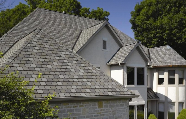A large, modern house in Madison WI with multiple gable roofs covered in grey shingles. The house features white walls, several windows, and is surrounded by lush green trees and bushes. Recent exterior renovations highlight a prominent tower-like structure.