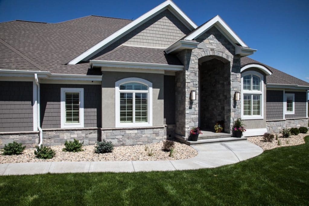 A modern single-story house in Madison, WI with a mix of stone and shingle exterior showcases large windows with white shutters and a prominent arched entrance. The neat front yard, recently improved by exterior renovations, has green grass, a curved pathway, and decorative plants near the entrance.