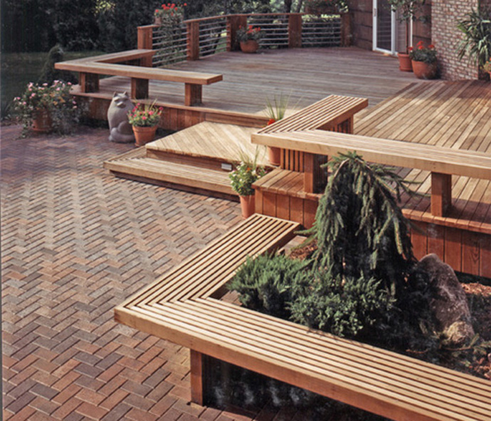 A spacious outdoor patio in Madison, WI features wooden decking with built-in benches. Steps connect brick flooring to the elevated deck. Potted plants and a small tree add greenery, while a stone figure of a cat decorates the area, showcasing recent exterior renovations.