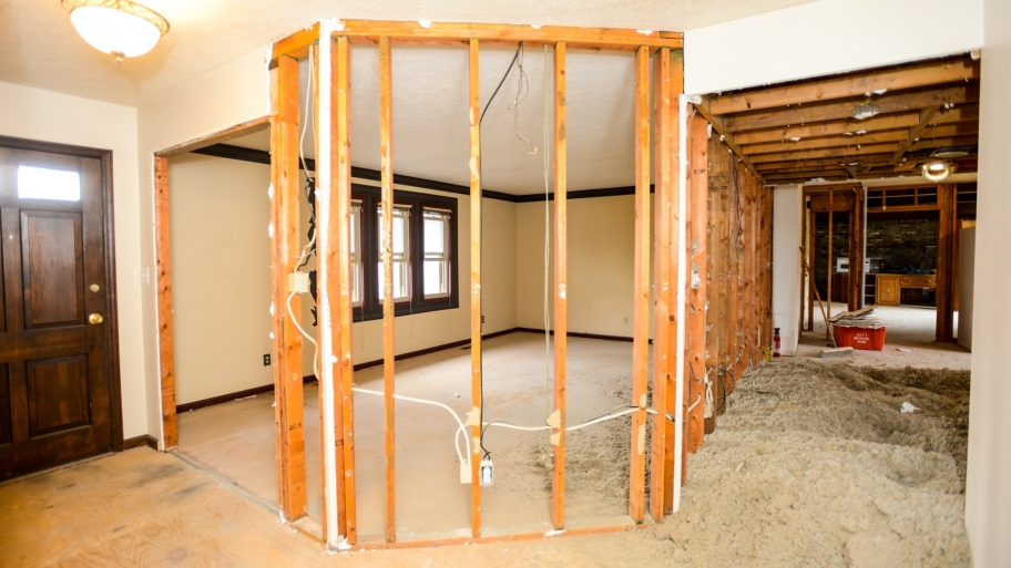 A house in Madison, WI, under renovation with exposed wooden framing and electrical wiring in the walls. The flooring is partially removed, revealing sand-like materials, and the ceiling structure is visible. There's a doorway leading to another room in the background, hinting at further exterior renovations.