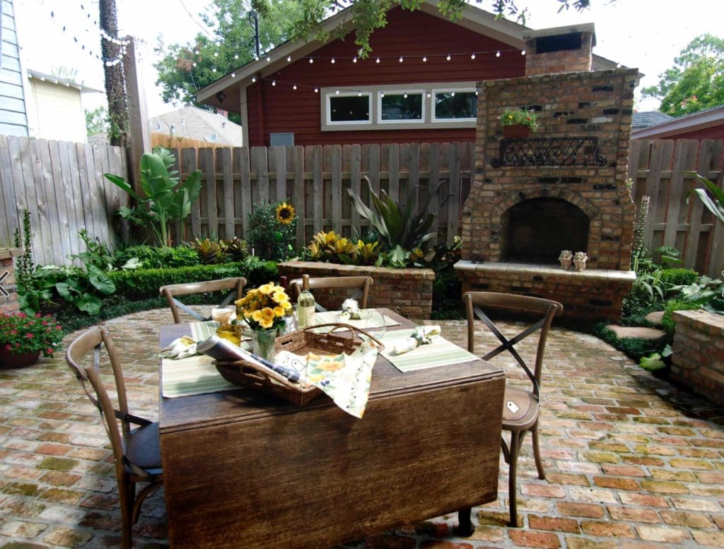 A cozy backyard patio with a wooden dining table set for four, featuring sunflowers and table settings. Located in Madison, WI, the area is paved with bricks and bordered by lush green plants. A brick outdoor fireplace stands at the back, with string lights hanging above—ideal for exterior renovations.