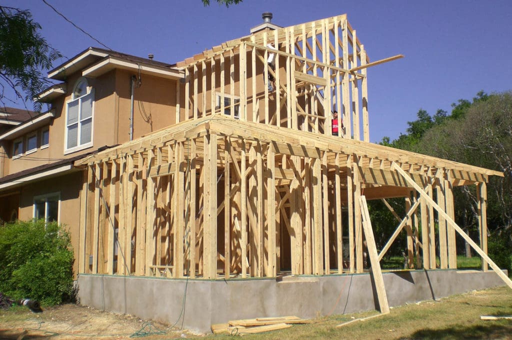 A residential house in Madison, WI with an ongoing exterior renovation project is shown. The house's beige exterior contrasts with the wooden frame structure being built as an extension. Planks, beams, and construction materials are scattered around, under a clear blue sky.