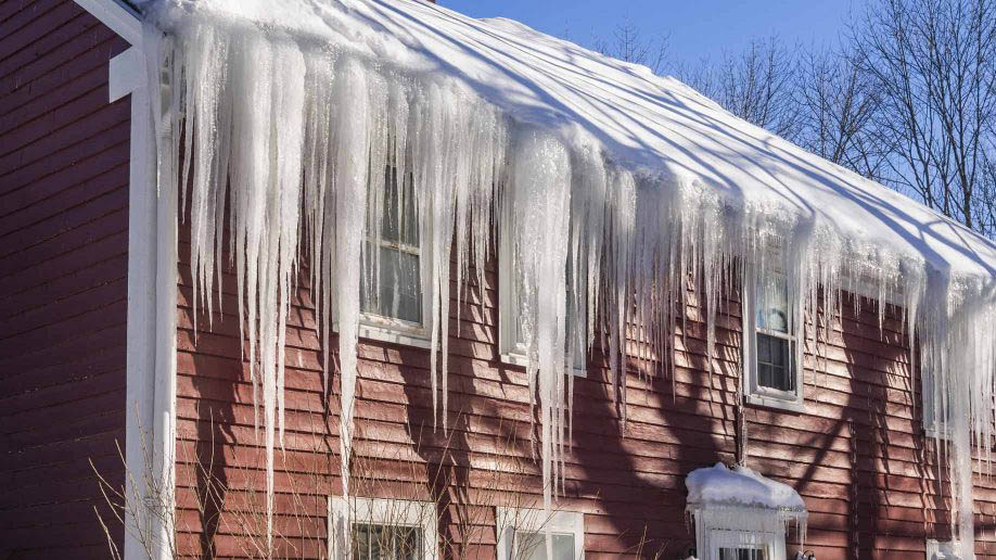 A red house in Madison, WI, with large icicles hanging from the snow-covered roof showcases recent exterior renovations. The sun is shining, casting shadows from the branches of nearby trees against a clear, blue sky. Several windows are visible on the house's side.