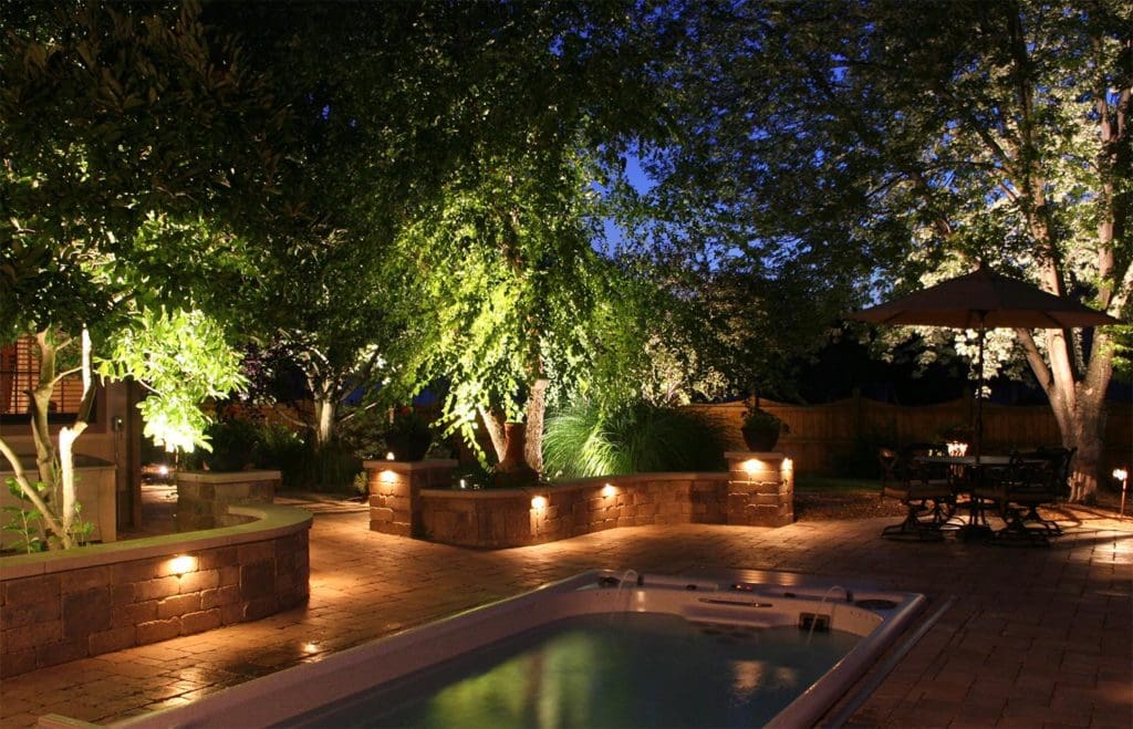A cozy backyard at dusk in Madison, WI, featuring a lit swimming pool, trees, and shrubs illuminated by ground lights. The paved patio area includes a seating arrangement under a large umbrella. Soft, warm lighting from recent exterior renovations creates a serene and inviting atmosphere.