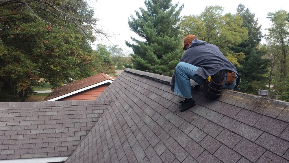 A person wearing a gray hoodie, blue jeans, and a brown cap is kneeling on a roof in Madison, WI while working on shingles. The individual has a tool belt with various tools. Trees and rooftops of nearby houses are visible in the background, highlighting the exterior renovations underway.