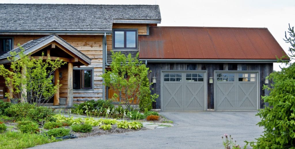 A rustic wooden house in Madison, WI, boasts a large two-door garage featuring grey doors. The exterior renovations include a blend of wood and dark metal siding with a steep, shingled roof. Out front, the well-maintained garden showcases various bushes, trees, and greenery along the paved driveway.