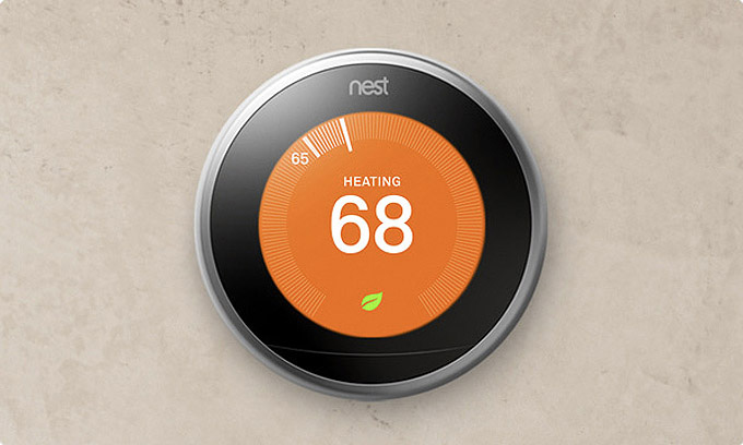 A Nest thermostat mounted on a wall in Madison, WI displays an orange screen with the text "HEATING" and the temperature set at 68°F. A small green leaf symbol is visible at the bottom of the screen. The sleek, round design with a silver border complements any recent exterior renovations.