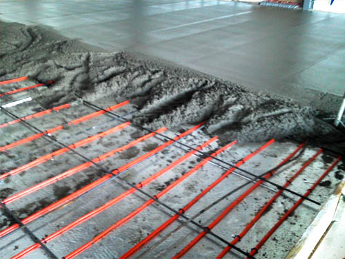 Concrete is being poured over a network of red heating tubes arranged in a grid pattern. The tubes are secured to a concrete subfloor with plastic clips. The fresh concrete is spreading evenly, partially covering the tubing, creating a new floor slab as part of ongoing exterior renovations in Madison, WI.