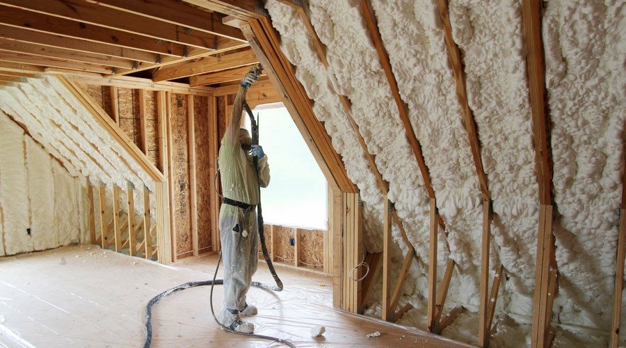 A person wearing protective gear sprays foam insulation on the walls of an unfinished attic space in a home in Madison, WI. The wooden frame of the roof is visible, and the floor is covered with a tarp to catch excess foam material. Natural light enters through a window in the background, highlighting ongoing exterior renovations.