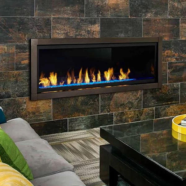 A modern, rectangular gas fireplace is built into a dark, tiled wall. Flames dance over a bed of blue and orange glass rocks. A glass coffee table with a yellow fruit bowl and a gray sofa with green and yellow cushions are partially visible in the foreground, perfect for cozy evenings after your exterior renovations in Madison, WI.