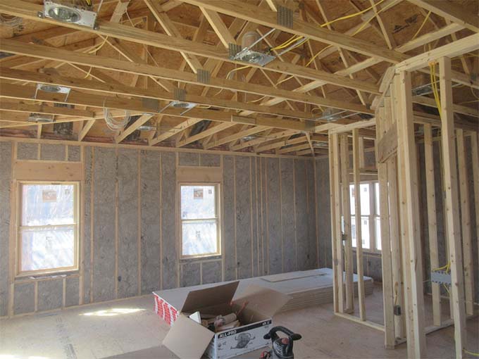 An unfinished room under construction in Madison, WI, with exposed wooden studs and rafters. Insulation partially covers the walls, and there are two windows. Various construction materials and tools are scattered on the floor, including a box of supplies and a vacuum cleaner, hinting at ongoing exterior renovations.