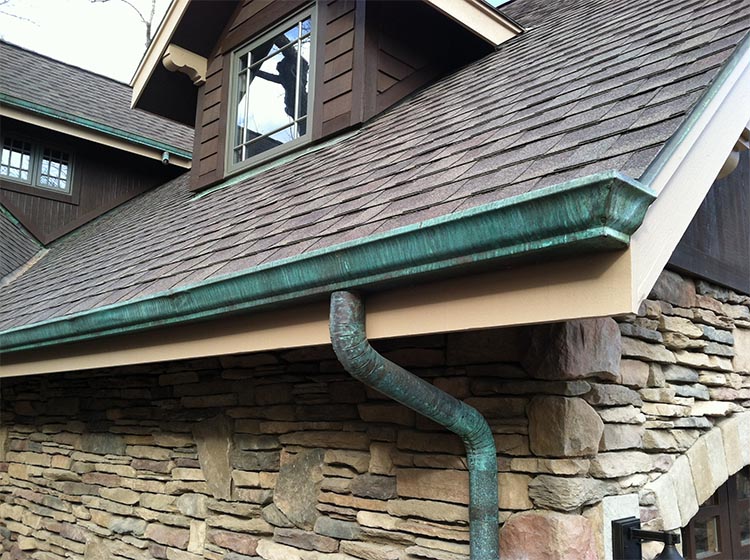 A slanted roof with brown shingles and weathered green copper gutters graces this Madison, WI home. The exterior wall is adorned with stone bricks, reflecting meticulous exterior renovations, while a portion of a gable window with a small roof is visible in the upper section of the image.