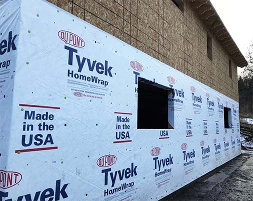 A building under construction in Madison, WI, is covered with Tyvek HomeWrap, displaying logos and text stating "DuPont Tyvek HomeWrap" and "Made in the USA". The wall has a cutout for windows, and some wooden framing is visible behind the wrap during exterior renovations.