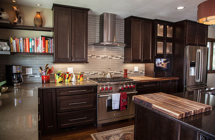 A modern kitchen in Madison, WI, features dark wood cabinets, stainless steel appliances, and a central island with a wooden countertop. Gray subway tile backsplash and colorful knives are displayed next to the stove, while cookbooks are neatly arranged on a shelf above the counter. Exterior renovations complement this stylish interior.