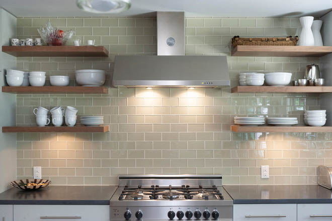 A modern kitchen in Madison, WI, with a stainless steel stove and vent. The backsplash features light green subway tiles. Wooden floating shelves hold white dishes, bowls, and mugs. The dark stone countertops have various kitchen items placed neatly amidst recent exterior renovations.