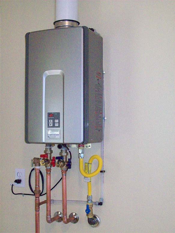 A wall-mounted tankless water heater with visible pipes and connections, including copper water lines and a yellow gas line, is an ideal choice for your exterior renovations in Madison, WI. The unit features control buttons and a digital display on the front, with an electrical outlet conveniently located to the left.