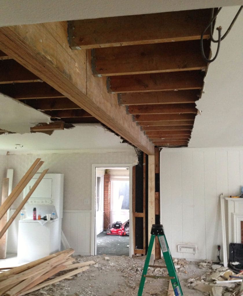 A room under renovation in Madison, WI, with exposed wooden beams and a partially demolished ceiling. Construction materials and debris are scattered on the floor. A green ladder is standing at the center, and there's a washing machine in the background, hinting at upcoming exterior renovations.