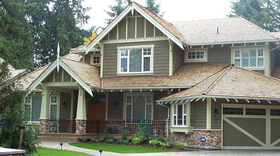 A two-story house in Madison, WI, features a green exterior with white trim and a wooden shingle roof. The front boasts large windows, a porch with stone pillars, and a landscaped yard. Recent exterior renovations include an attached garage on the right side of the house. Trees are visible in the background.