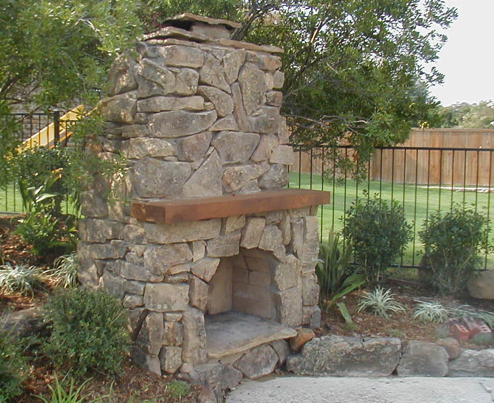 An outdoor stone fireplace with a wooden mantel stands surrounded by greenery, including bushes and trees, in this elegant Madison, WI retreat. In the background, a black metal fence and a wooden privacy fence enclose a grassy yard, showcasing the meticulous exterior renovations.