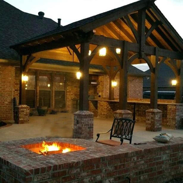 A cozy outdoor patio in Madison, WI, features a brick fire pit with a blazing fire, surrounded by brick pillars. The area is illuminated by warm lights hanging from a wooden pergola roof. A decorative metal chair is placed nearby, and a brick house showcasing recent exterior renovations is visible in the background.