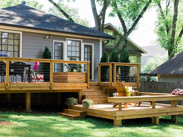 A cozy backyard in Madison, WI with a wooden deck featuring a multi-level design. There are potted plants, a table with chairs on one side, and a lower level with armchairs and a table. The house has dark gray siding and French doors leading to the deck. Several trees provide shade—perfect for exterior renovations.