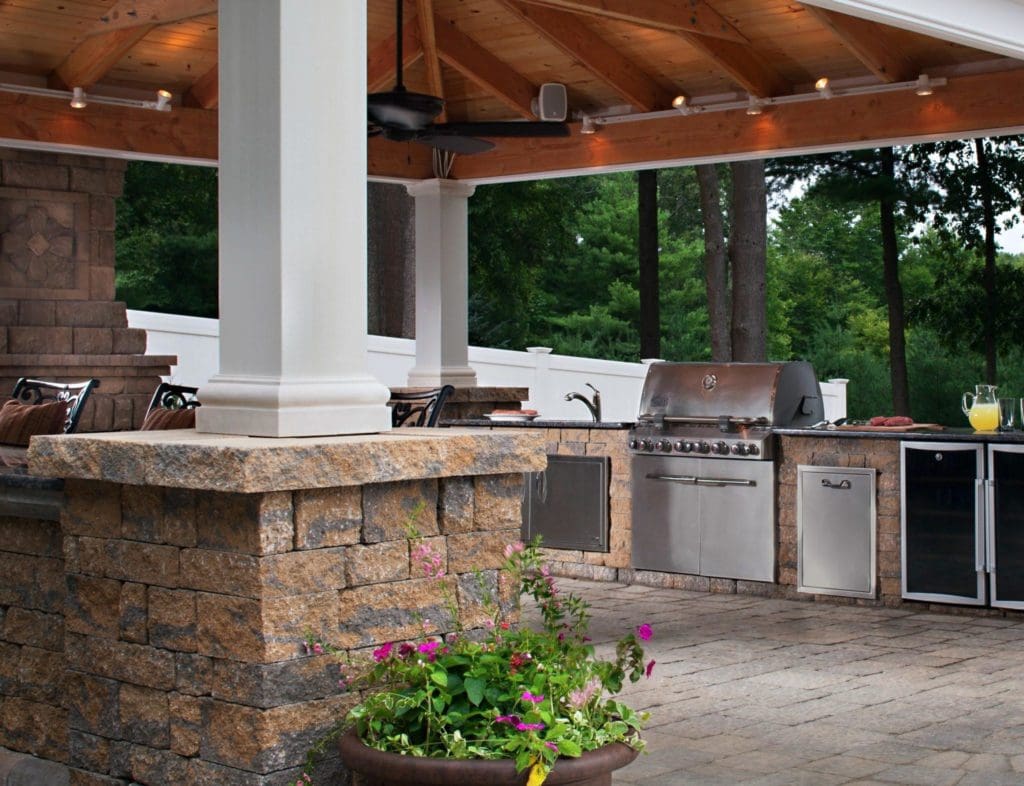 Outdoor kitchen with stone countertops and stainless steel appliances, including a grill and refrigerator, under a wooden pergola with lighting. A planter with colorful flowers is in the foreground, enhancing this exterior renovation. Trees provide a scenic backdrop in Madison, WI.