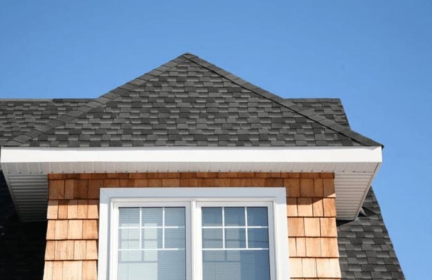 A close-up of a house's exterior in Madison, WI, showing a section of the roof with dark gray asphalt shingles, a white-trimmed peak, and cedar shake siding. Below the roof peak, there are two white-framed windows with grids and blinds against a blue sky backdrop—perfect inspiration for exterior renovations.