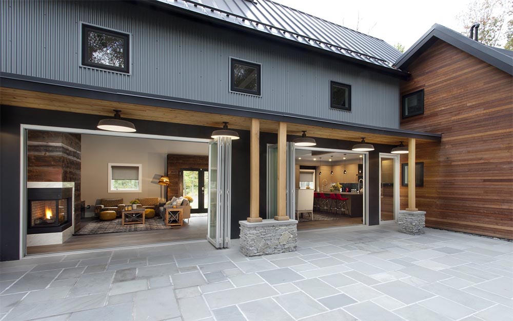 A modern house with gray and wooden exterior renovations. The open outdoor space reveals a cozy living room with a fireplace and a kitchen, both visible through large, folding glass doors. Located in Madison, WI, the patio is paved with gray stones and has a rustic touch with wooden beams and stone bases.