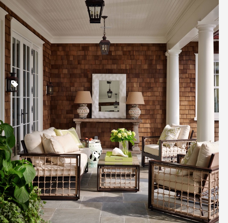 A cozy outdoor patio in Madison, WI features a beige couch and two matching armchairs with rope detailing. A square coffee table with a vase of green flowers and a white ceramic stool sits in the center. A textured wooden wall, white columns, and hanging lanterns complete this stunning exterior renovation.