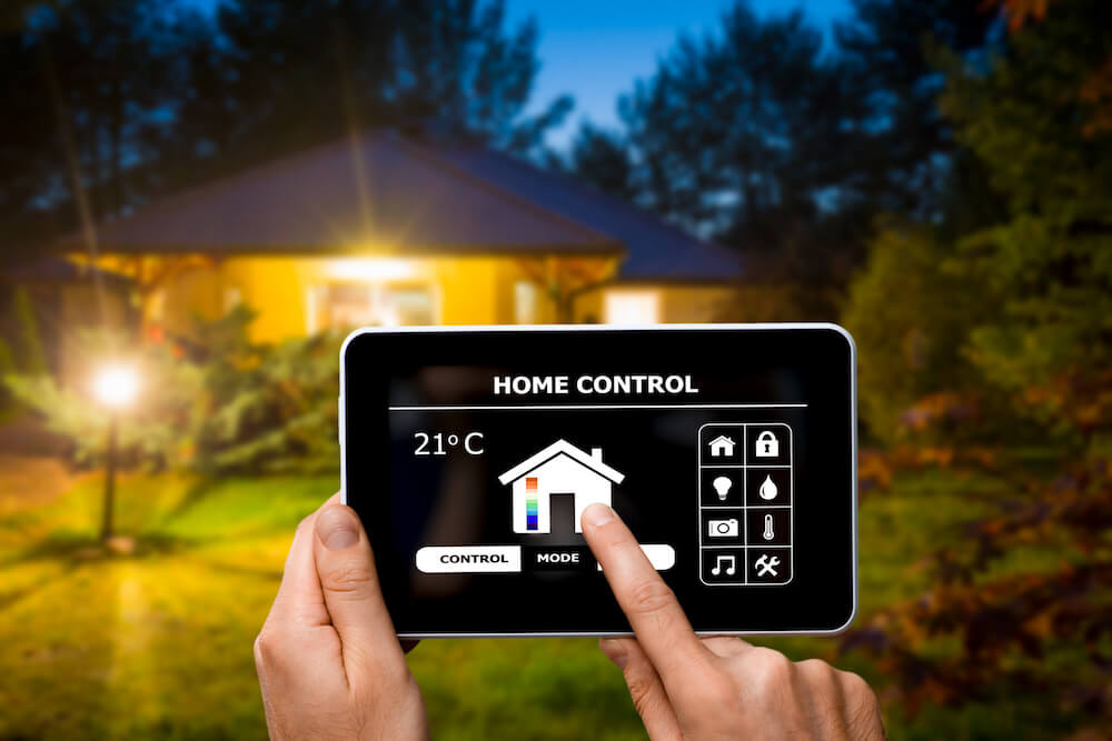 A person in Madison, WI holds a tablet displaying a home automation interface labeled "Home Control." Various icons for controlling temperature, lights, security, and other household functions are visible on the screen. A house undergoing exterior renovations is illuminated in the background at dusk.