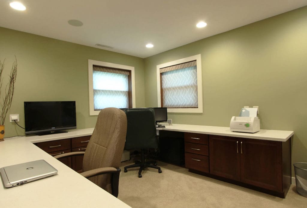A home office in Madison, WI with sage green walls features a large L-shaped desk with wooden cabinets and white countertops. Two office chairs face desktop computers. A printer is on the right, and a closed laptop is on the left counter. Two windows with translucent blinds open to views of recent exterior renovations.