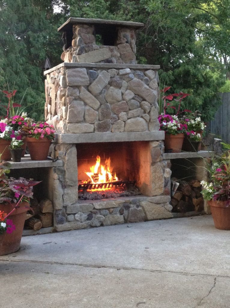 A stone outdoor fireplace burns with a bright, warm fire. It is surrounded by potted plants filled with colorful flowers. A small stack of firewood is neatly piled to the right of the fireplace. In the background, green trees provide a lush, natural setting—ideal for exterior renovations in Madison, WI.
