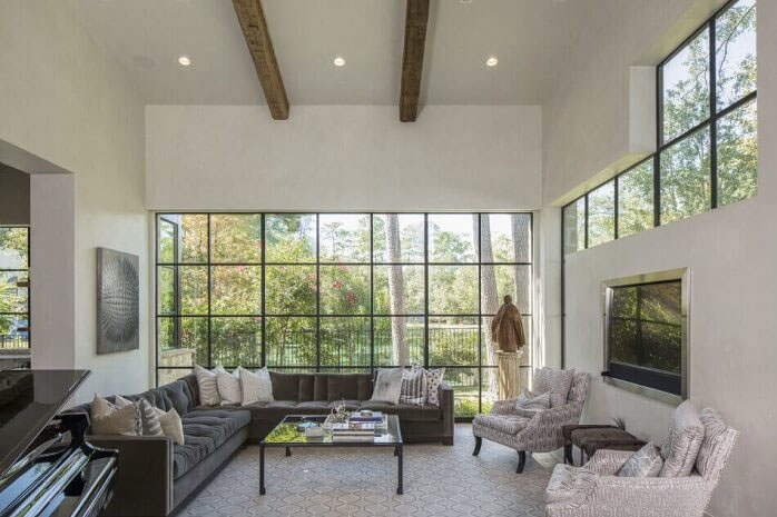 A spacious, modern living room in Madison, WI with high ceilings and large windows allowing natural light to flood the space. The room features a gray sectional sofa, two patterned armchairs, a glass coffee table, and a wall-mounted TV. Decorative pillows and greenery add warmth while exterior renovations enhance its charm.