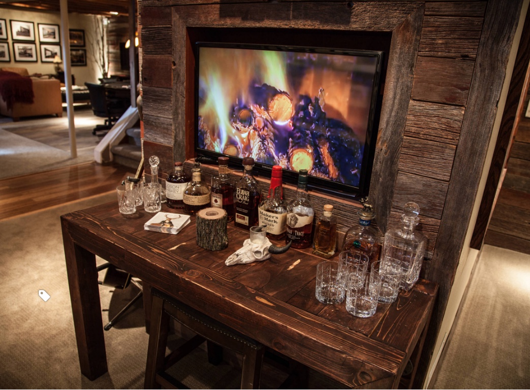 A cozy room in Madison, WI features a wooden table with an assortment of liquor bottles, glasses, and bar accessories. Behind the table is a flatscreen TV displaying a crackling fire set in a rustic wooden wall. The space exudes a warm, inviting ambiance perfect for relaxing after exterior renovations.