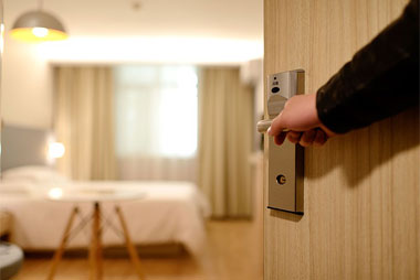 An open door reveals a well-lit hotel room in Madison, WI, showing a bed with white linens, a small round table, and curtains covering a large window. A hand is holding the door handle, suggesting someone is entering the room amidst ongoing exterior renovations.