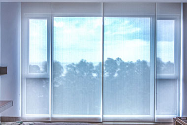 A large window with sheer white curtains partially drawn allows a view of a serene, cloudy sky and tree silhouettes outside, illustrating the charm of Madison, WI. The room has minimal visible furnishings, creating a calm and airy atmosphere ideal for contemplating exterior renovations.