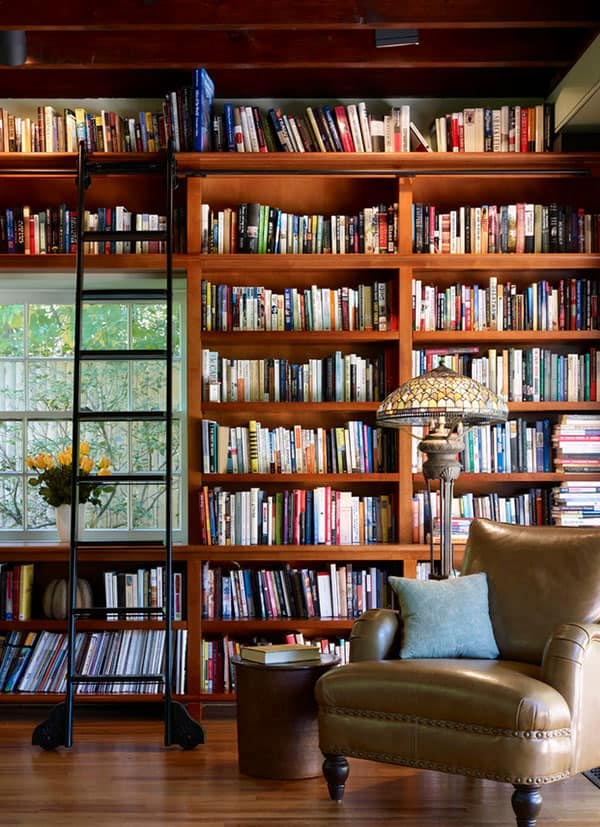 A cozy reading nook in Madison, WI, featuring tall wooden bookshelves filled with books, a ladder on the left, a window with greenery outside from recent exterior renovations, an antique table lamp with a stained glass shade, a leather armchair with a blue pillow, and a small round wooden side table.