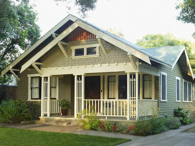 A charming single-story house in Madison, WI, featuring a green exterior and white trim. The home boasts a front porch with a gabled roof, white railings, and decorative support beams. Exterior renovations include lush greenery and flower beds that surround the pathway leading to the front door.