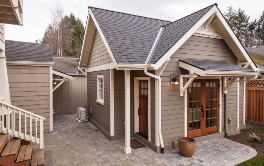 A charming, small house in Madison, WI with gray siding and white trim features a wooden front door with large glass panels and a sloped roof. The exterior renovations also include a paved walkway and a potted plant by the entrance.