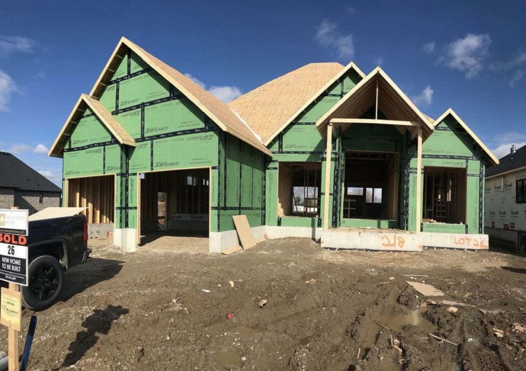 A partially constructed house with a green exterior is shown on a sunny day in Madison, WI. The wooden framing is visible, and the roof is incomplete. A "Sold" sign stands in front, and various construction materials are scattered around the muddy front yard, indicating ongoing exterior renovations.