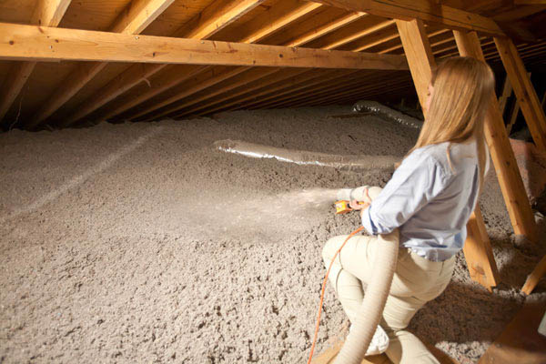 A person in a light blue shirt and beige pants is kneeling in an attic with wooden beams and a pitched roof, using a hose to blow light grey insulation material onto the attic floor. As part of their exterior renovations in Madison, WI, this task ensures optimal energy efficiency for the home.