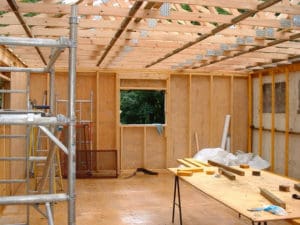 Interior view of a wooden framed construction site with an unfinished ceiling. Scaffolding is on the left, tools and wooden planks are on a table in the center, and building materials are scattered around. A window opening offers a glimpse of the greenery outside, typical of many exterior renovations in Madison WI.