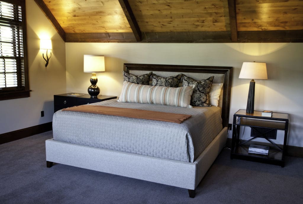 A cozy bedroom with a sloped wooden ceiling features a neatly made bed with patterned pillows and a light gray comforter. Two bedside tables, each with a lamp, flank the bed. A window with wooden blinds is on the left, and a wall sconce light adds additional warmth. Perfect for unwinding after busy days tackling exterior renovations in Madison, WI.