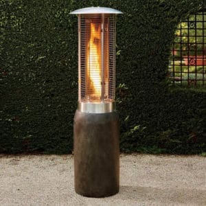 Tall patio heater with a cylindrical stainless-steel body and a protective metal grill. Flames are visible inside the heater, providing warmth. This elegant addition to your outdoor space is perfect for exterior renovations, situated on a gravel path with a green hedge and wrought iron gated window behind.