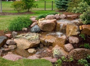 A landscaped garden in Madison, WI features a small waterfall cascading over large, natural rocks surrounded by greenery, bushes, and mulch. In the background, a metal fence and more green foliage complete this serene outdoor scene, perfect for showcasing your exterior renovations.