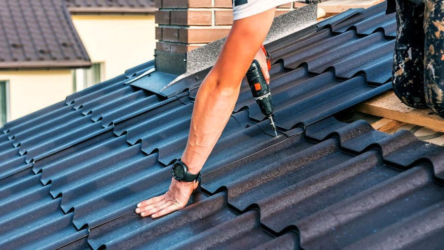 A person using a power drill to secure a metal roof panel during exterior renovations. Their left hand stabilizes the panel while their right hand operates the drill. A brick chimney is partially visible, and additional roof panels and structures are in the background, showcasing work in Madison, WI.