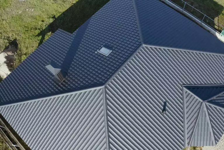 Aerial view of a house’s roof in Madison, WI, showcasing dark grey corrugated metal roofing. The exterior renovations include several sections with varying slopes, a skylight near the center, and a small chimney. The surrounding area comprises a grassy lawn and partially visible scaffolding.