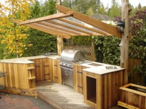 Outdoor kitchen with wooden countertops and cabinetry, featuring a stainless steel grill, sink, and ample counter space. The setup is under a transparent pergola. The area is surrounded by lush greenery in Madison, WI making it a cozy and functional cooking space for exterior renovations.